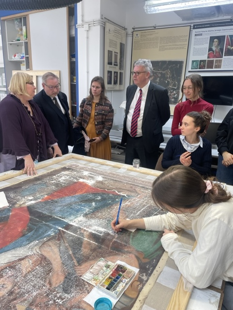 kobieta w okularach tłumaczy grupie osób zabiegi konserwatorskie. Na pierwszym planie młoda dziewczyna retuszuje obraz farbami