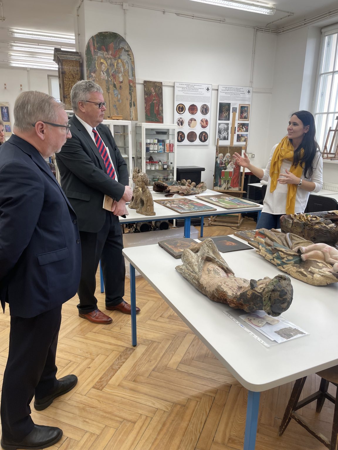 kobieta w białej bluzce i żółtym szaliku tłumaczy zabiegi konserwatorskie dwóm mężczyznom
