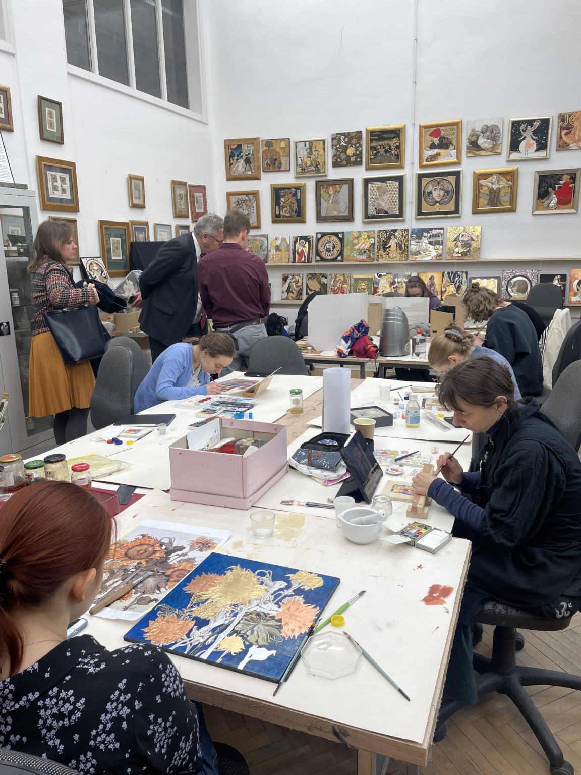 studenci na zajęciach z pozłotnictwa pracują przy stołach. W kącie pracowni mężczyźni obserwują prace studentów