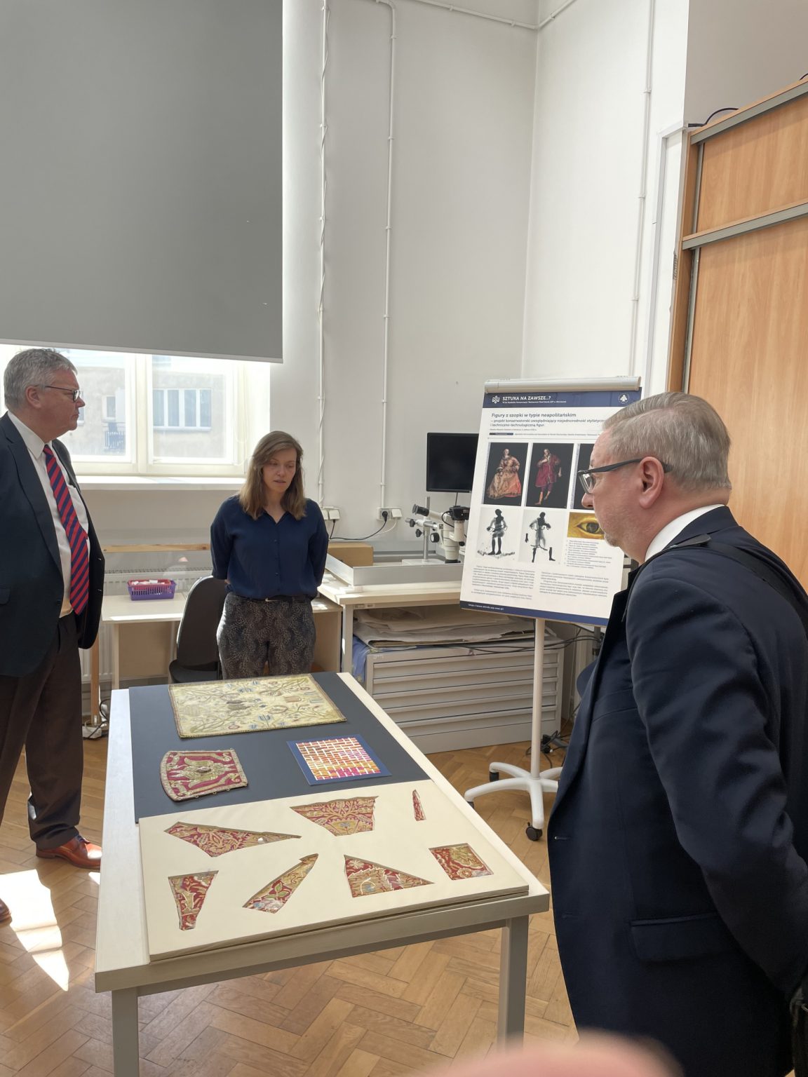 dwóch mężczyzn w garniturach i jedna kobieta stoją przy stole na którym leżą fragmenty tkanin