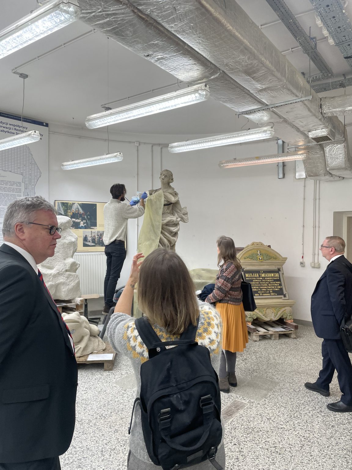 na pierwszym planie kobieta w szarym swetrze i z czarnym plecakiem. W pracowni grupa osób obserwuje konserwację rzeźby