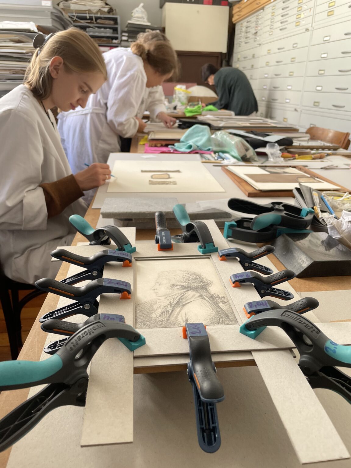 studentki ubrane w białe fartuchy pracują przy zabytkach papierowych. Na pierwszym planie obiekt unieruchomiony ściskami