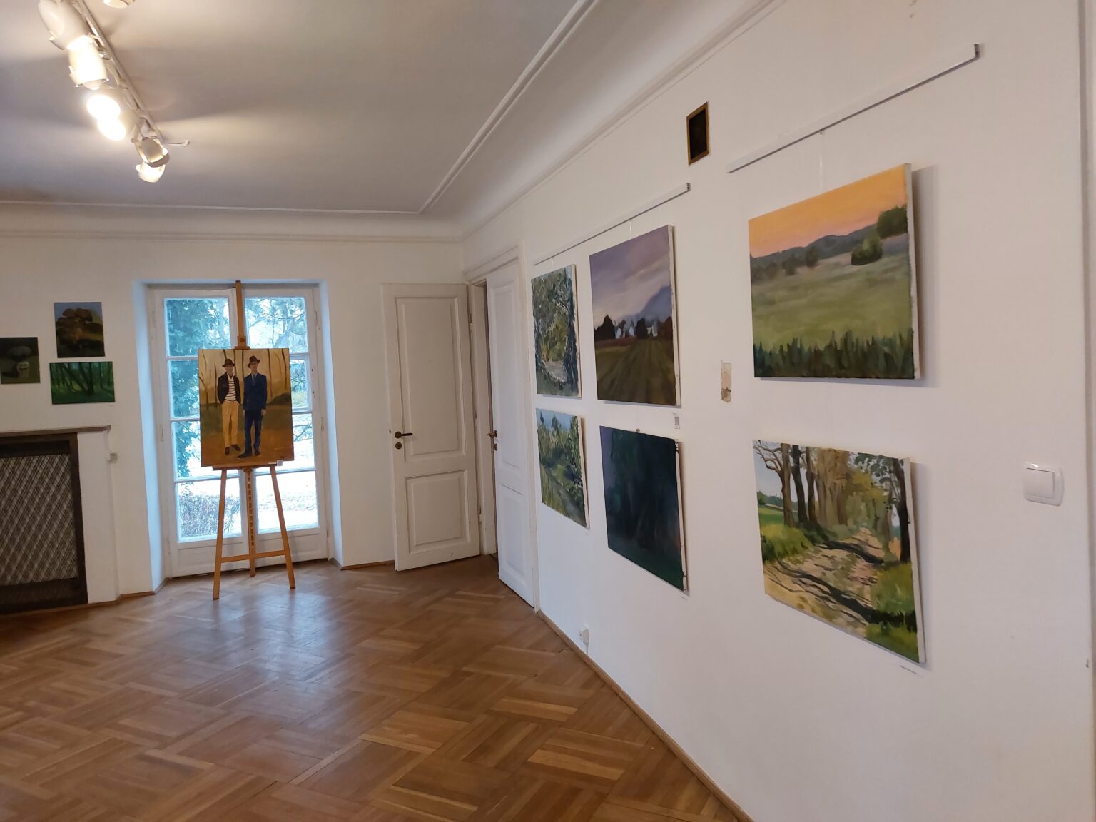 wystawa prac. Na białych ścianach widzą prace, Na wprost okna na sztaludze stoi obraz z postaciami dwóch mężczyzn