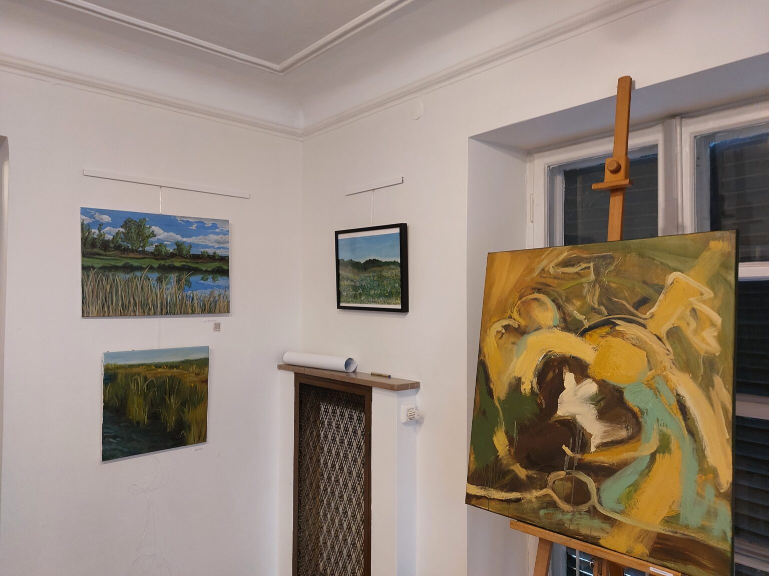 fragment wystawy, na ścianie wiszą 3 prace przedstawiające krajobrazy. Po prawej stronie na sztaludze stoi abstrakcyjny obraz w kolorach żółto zielonych
