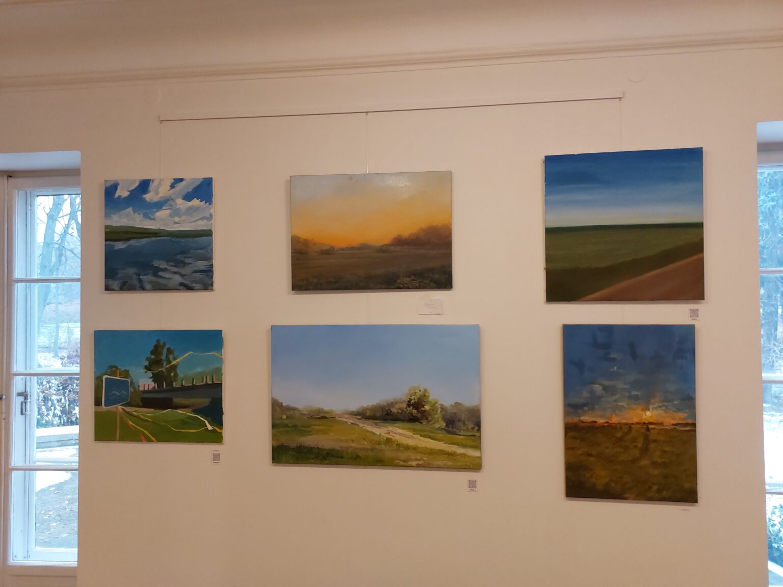 wystawa prac. Na białej ścianie wisi sześć prac przedstawiających pejzaże, krajobraz z łąkami i i błękitnym niebem