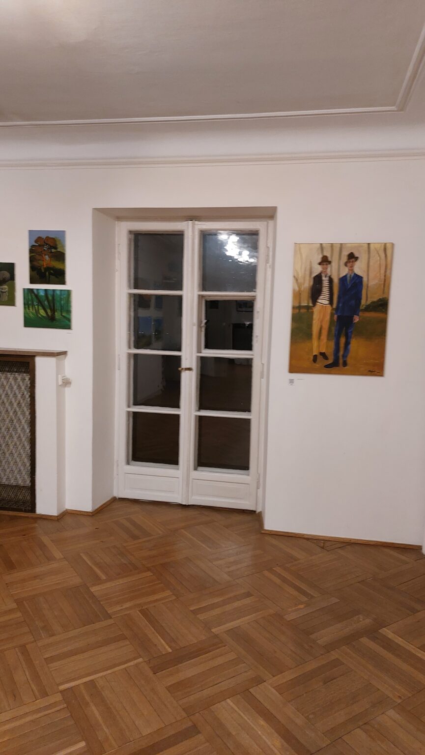 wystawa prac. Na środku zdjęcia okno balkonowe, po bokach wiszą prace na tle białych ścian