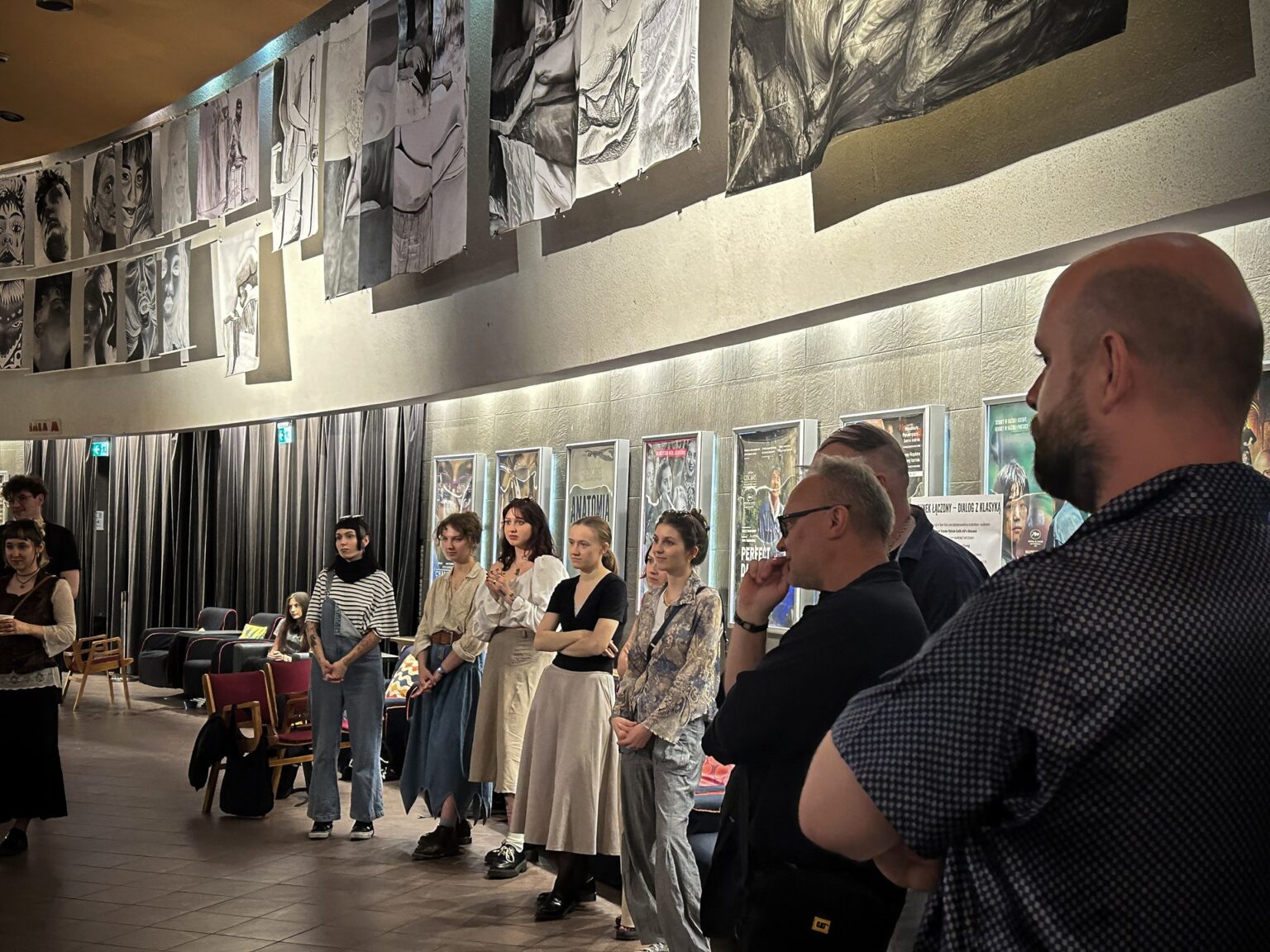 Grupa osób stoi w pomieszczeniu o charakterze galerii lub przestrzeni wystawienniczej, gdzie prezentowane są dzieła sztuki lub fotografie.