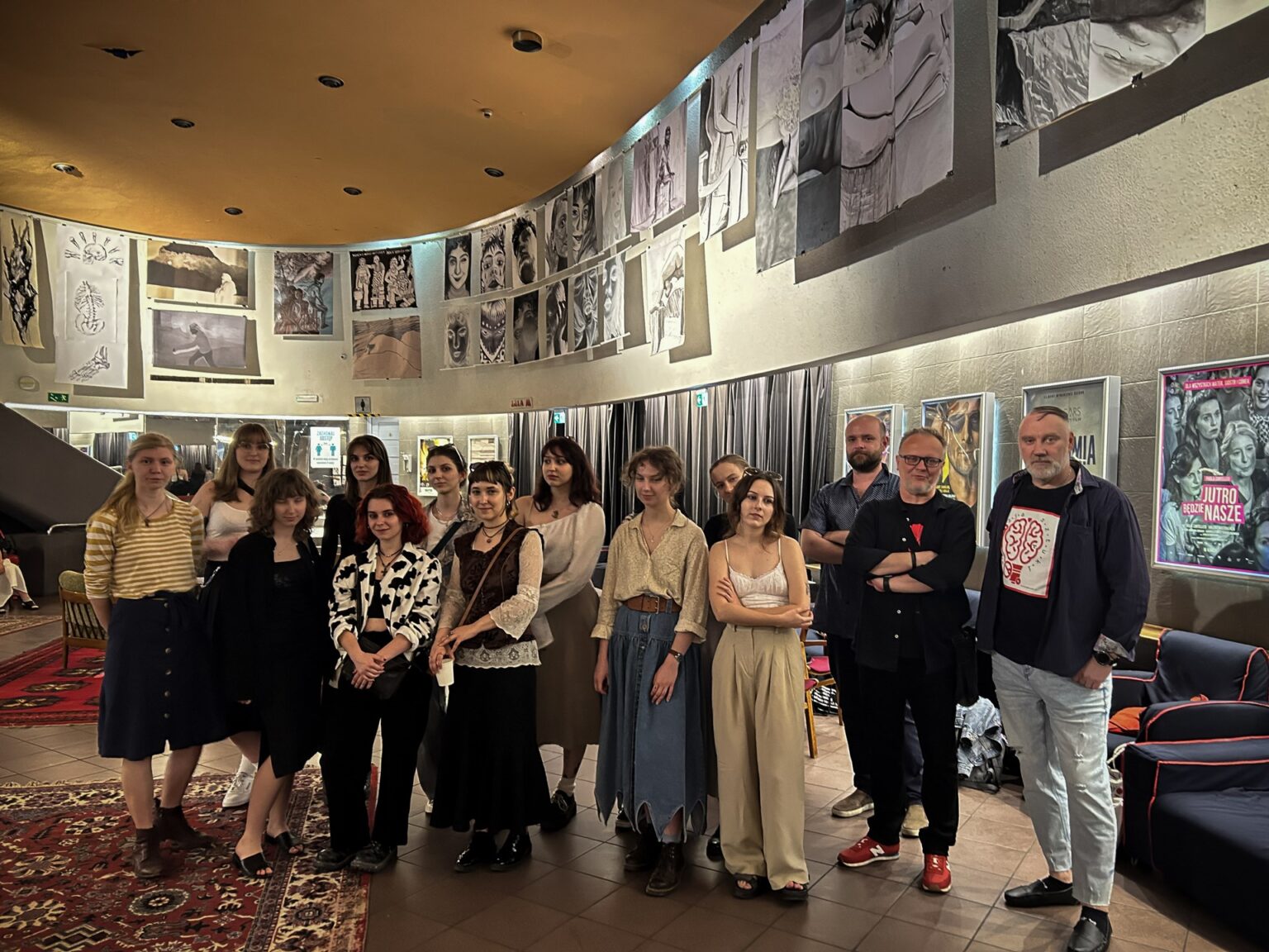 Grupa osób stoi w pomieszczeniu o charakterze galerii lub przestrzeni wystawienniczej, gdzie prezentowane są dzieła sztuki lub fotografie.