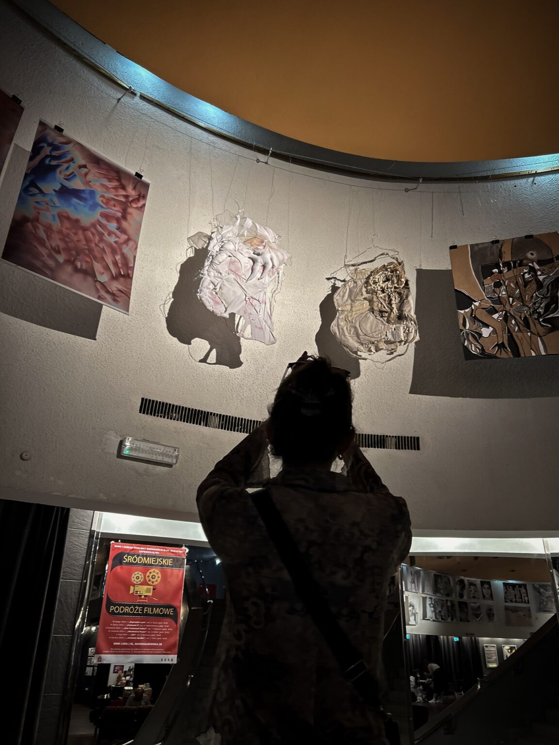 osoba stoi w pomieszczeniu o charakterze galerii lub przestrzeni wystawienniczej, gdzie prezentowane są dzieła sztuki lub fotografie.