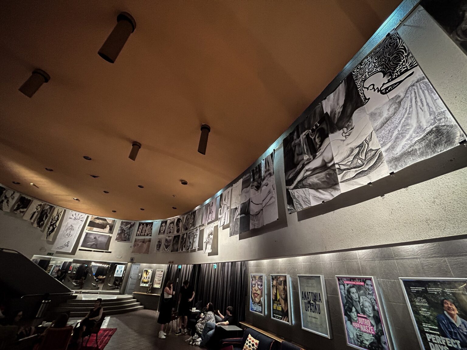 Grupa osób stoi w pomieszczeniu o charakterze galerii lub przestrzeni wystawienniczej, gdzie prezentowane są dzieła sztuki lub fotografie.