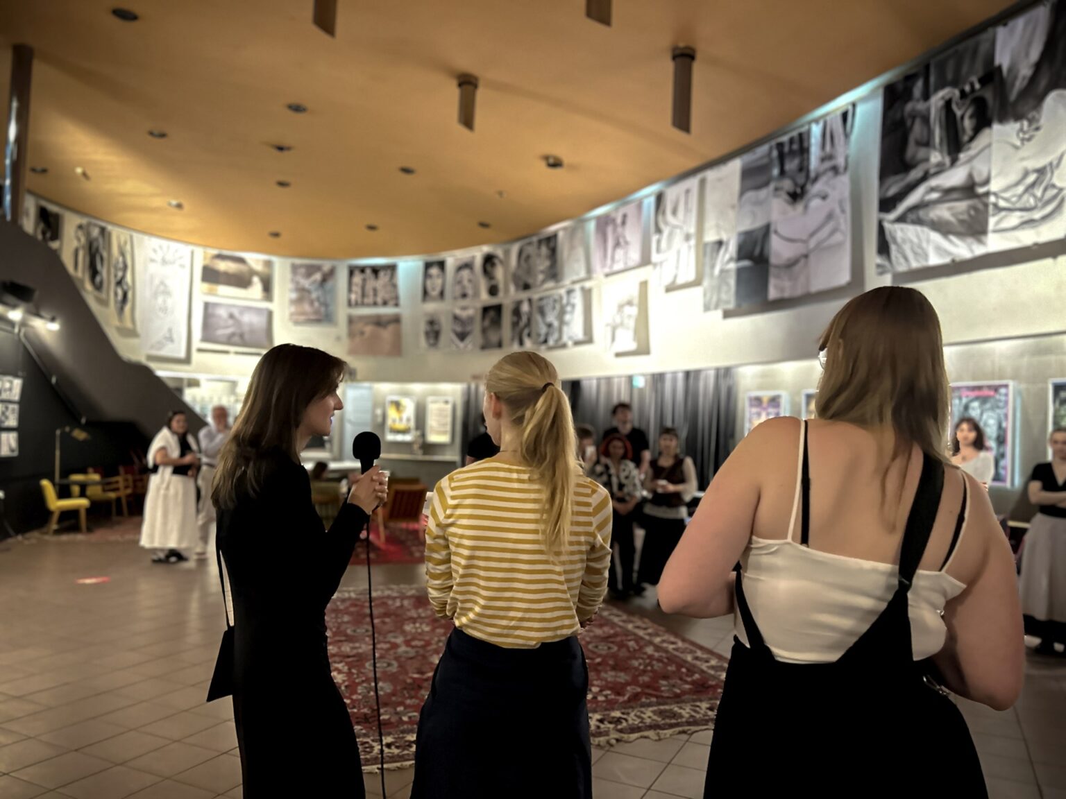 Grupa osób stoi w pomieszczeniu o charakterze galerii lub przestrzeni wystawienniczej, gdzie prezentowane są dzieła sztuki lub fotografie.