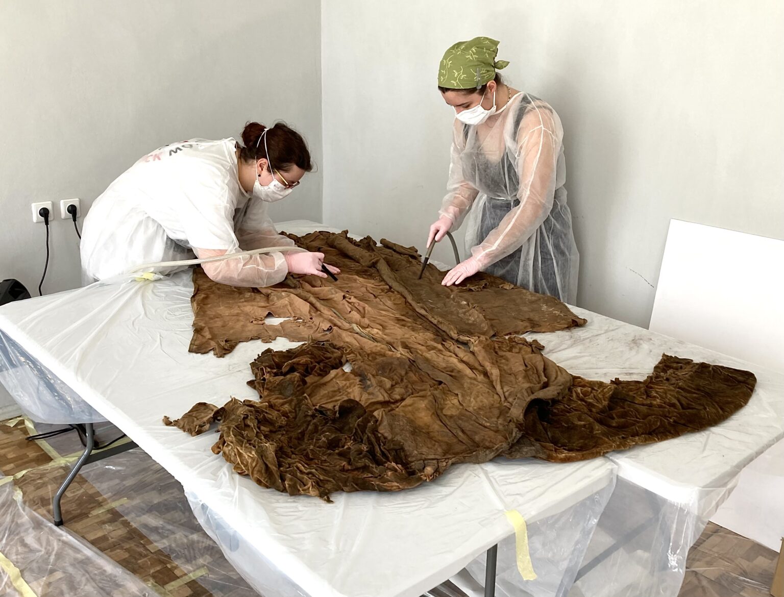 dwie osoby ubrane w strój ochrony nachyla się na tkaniną leżącą na stole