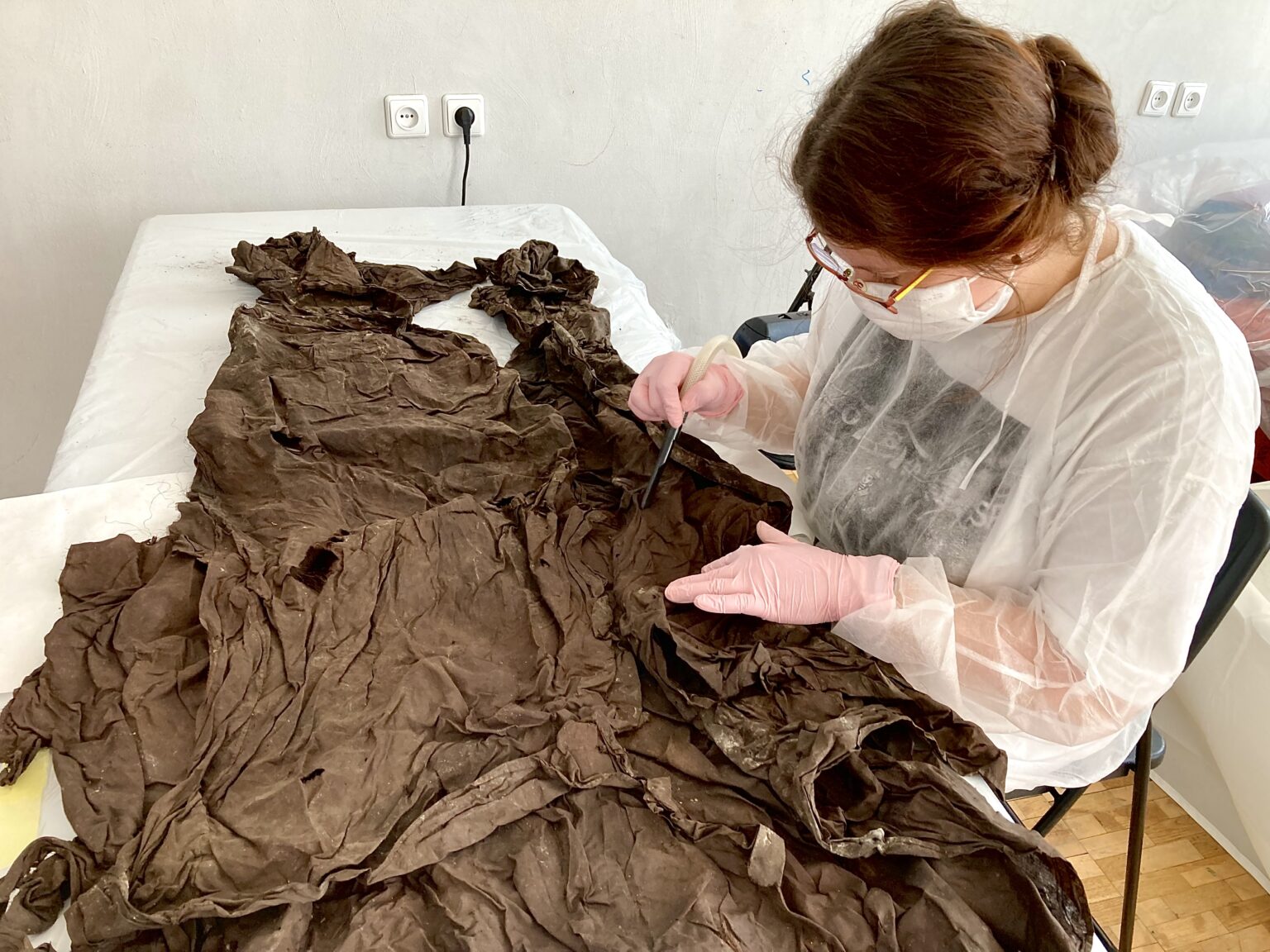 osoba ubrana w biały strój ochrony nachyla się na tkaniną leżącą na stole