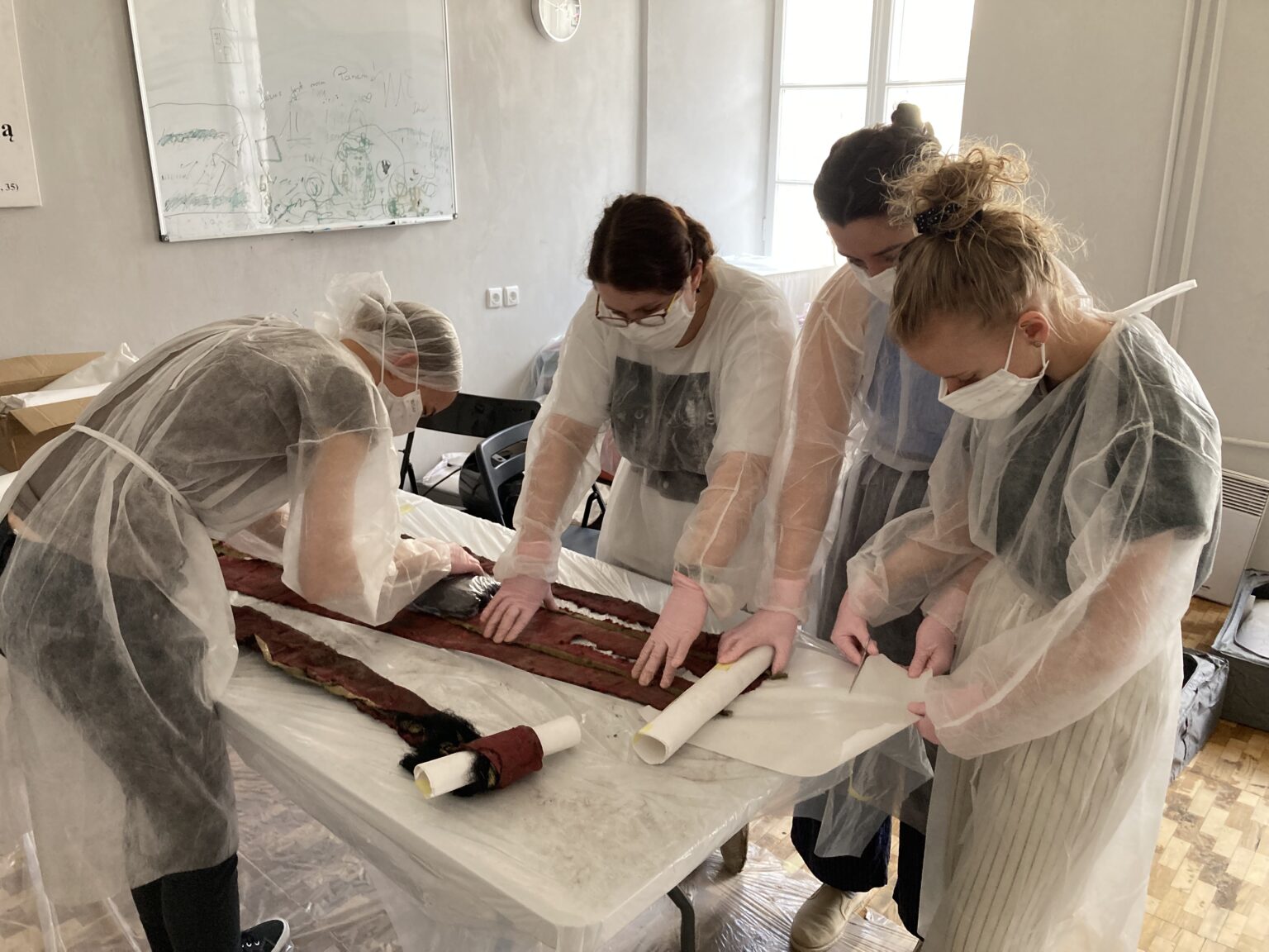 cztery osoby ubrane w strój ochrony nachyla się na tkaniną leżącą na stole