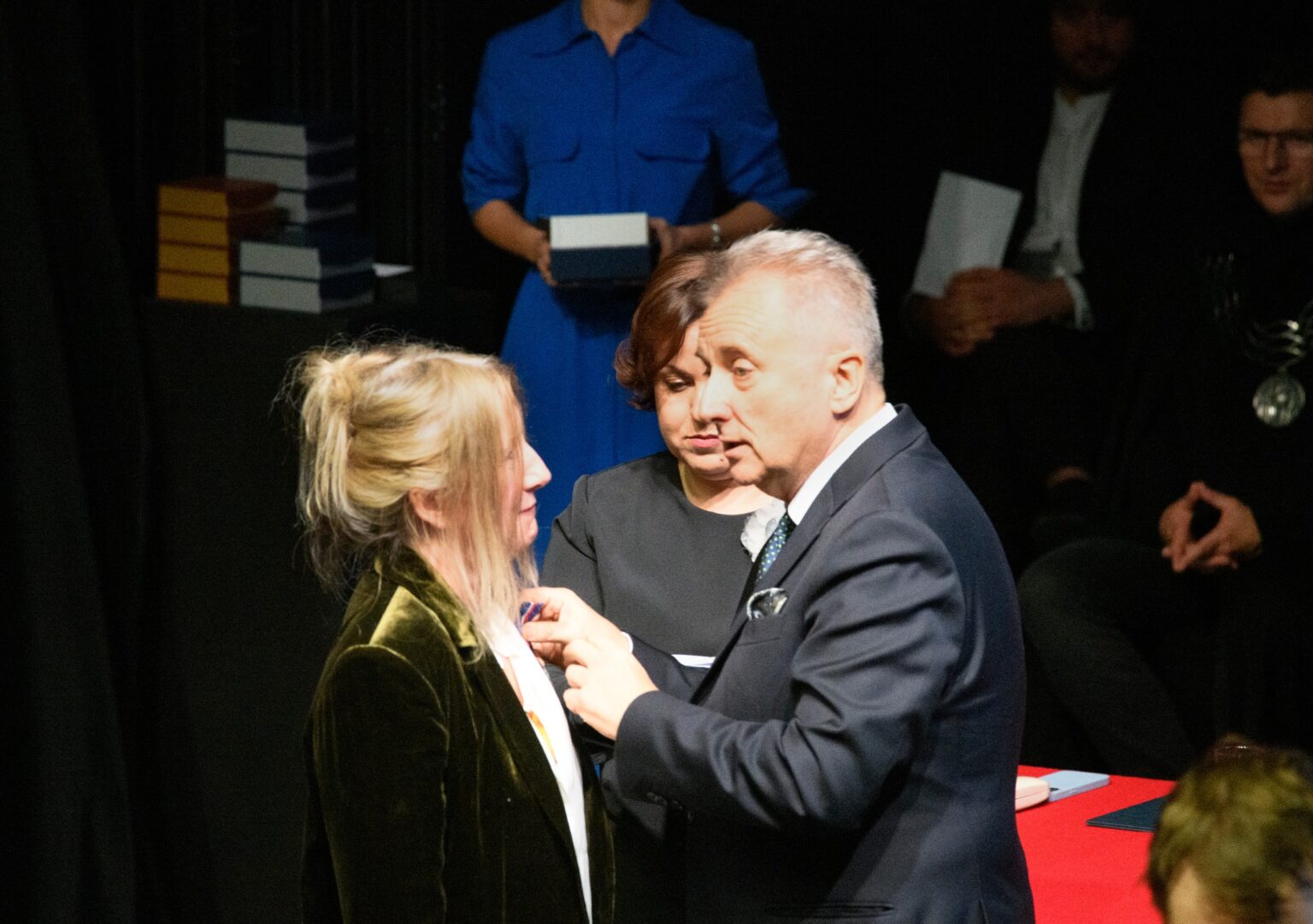 Mężczyzna w garniturze przypina medal kobiecie o blond włosach, ubranej w ciemno szary żakiet