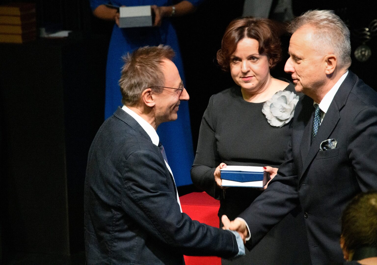 Dwóch mężczyzn ściska sobie dłonie, w tle kobieta ubrana na czarno trzymająca w rękach pudełko