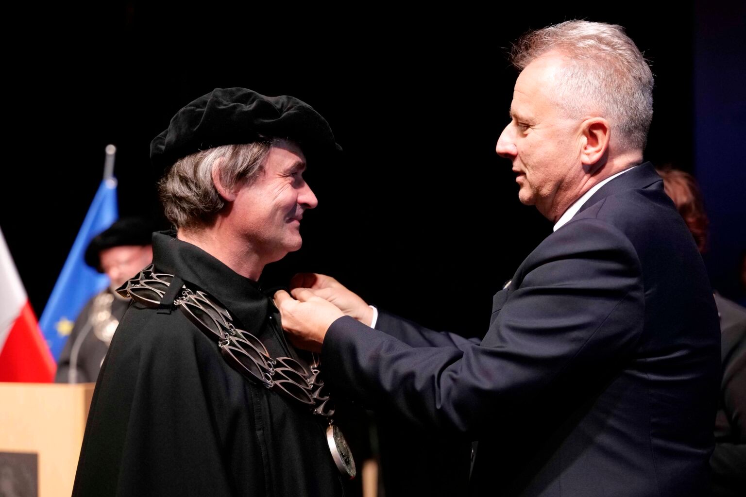 mężczyzna w garniturze przypina medal drugiemu mężczyźnie w czarnej todze.