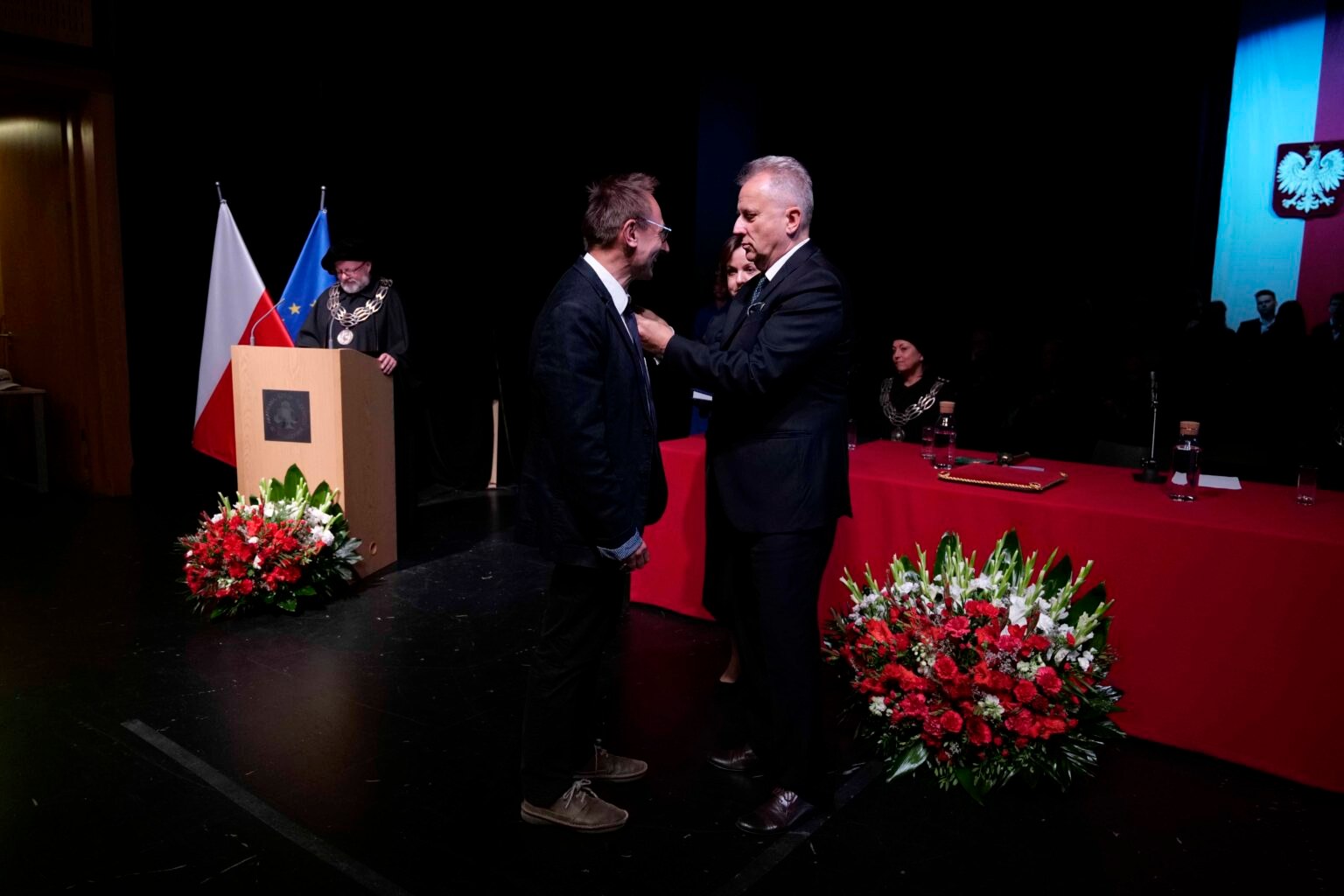 mężczyzna w garniturze przypina medal drugiemu mężczyźnie. Oboje ubrani są na czarno, w tle czerwony stół i kwiaty