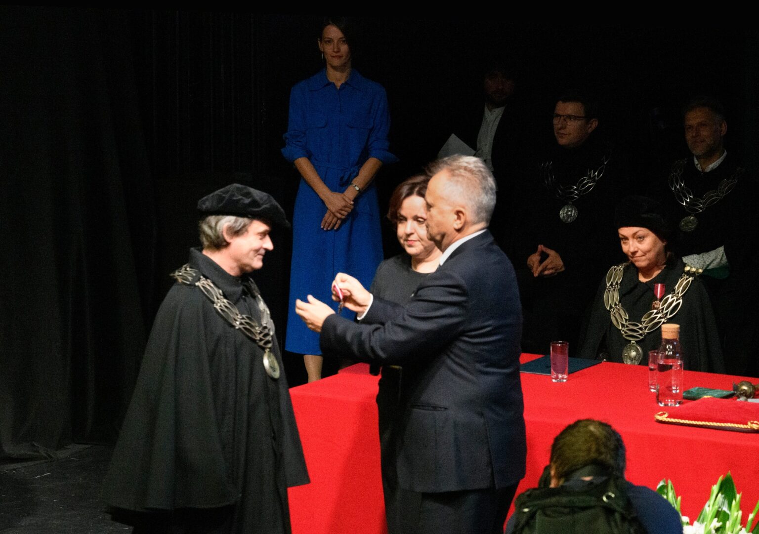 mężczyzna w garniturze przypina medal drugiemu mężczyźnie. Oboje ubrani są na czarno, w tle czerwony stół i kwiaty