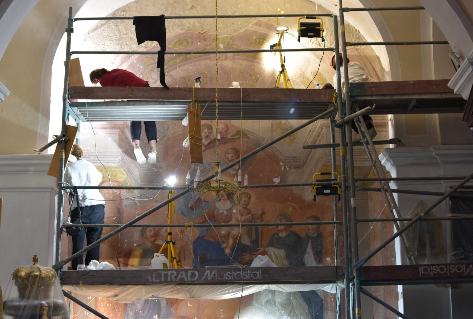 Na zdjęciu widzimy scenę wewnętrznych prac konserwatorskich. W centrum znajduje się duża konstrukcja rusztowania, na której pracują dwie osoby. Jedna z nich, ubrana w czerwoną koszulę i ciemne spodnie, stoi na górnym poziomie rusztowania i wydaje się pracować nad sufitem lub górną częścią ściany, która nie jest w pełni widoczna. Druga osoba stoi na niższym poziomie rusztowania i obserwuje lub czeka. W tle widoczna jest część ozdobnego malowidła przedstawiającego kilka postaci, co sugeruje, że może to być budynek historyczny lub religijny. Na platformach rusztowania rozłożone są różne narzędzia i sprzęt. W lewym dolnym rogu obrazu widać złotą statuę, częściowo zasłoniętą przez rusztowanie.