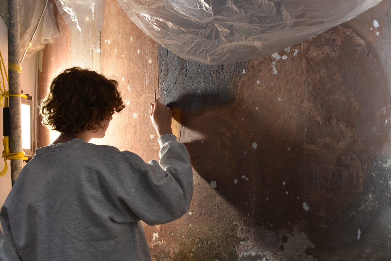 Na zdjęciu widzimy osobę malującą ścianę na ciemny kolor. Osoba ta ma na sobie szarą bluzę z długim rękawem i używa wałka malarskiego. Ściana jest w trakcie remontu, widać na niej niepomalowane fragmenty oraz białe plamy wypełniacza lub podkładu. Nad ścianą wisi folia ochronna, co sugeruje, że podjęto środki ostrożności, aby chronić otoczenie przed rozpryskami farby.