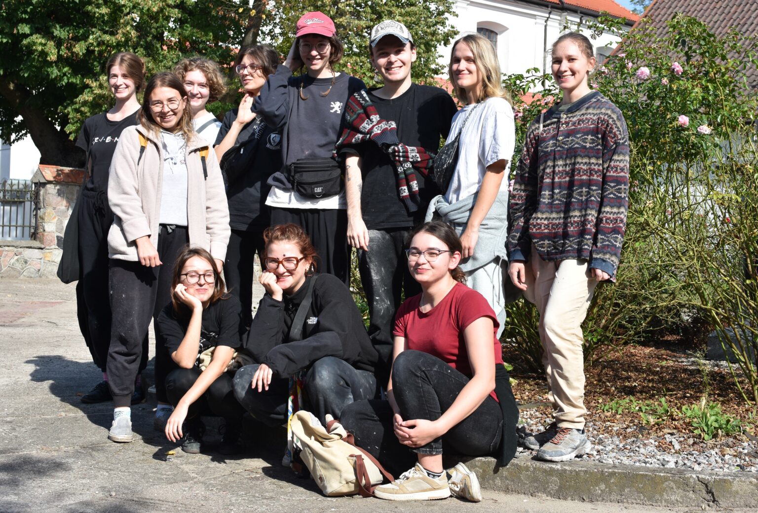 Na zdjęciu widzimy grupę osób stojących razem na zewnątrz, do zdjęcia grupowego. Widoczne są różne style ubioru, co sugeruje swobodną atmosferę.