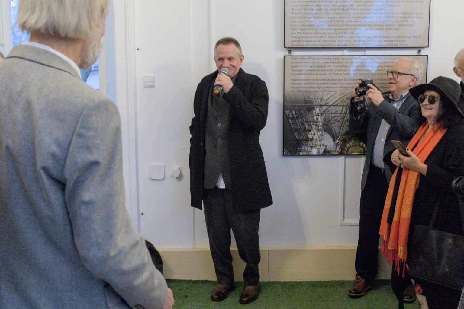 Mężczyzna z mikrofonem przemawia podczas wydarzenia, stojąc na tle plansz wystawowych. Wokół niego zgromadzeni są uczestnicy wydarzenia, część z nich robi zdjęcia.