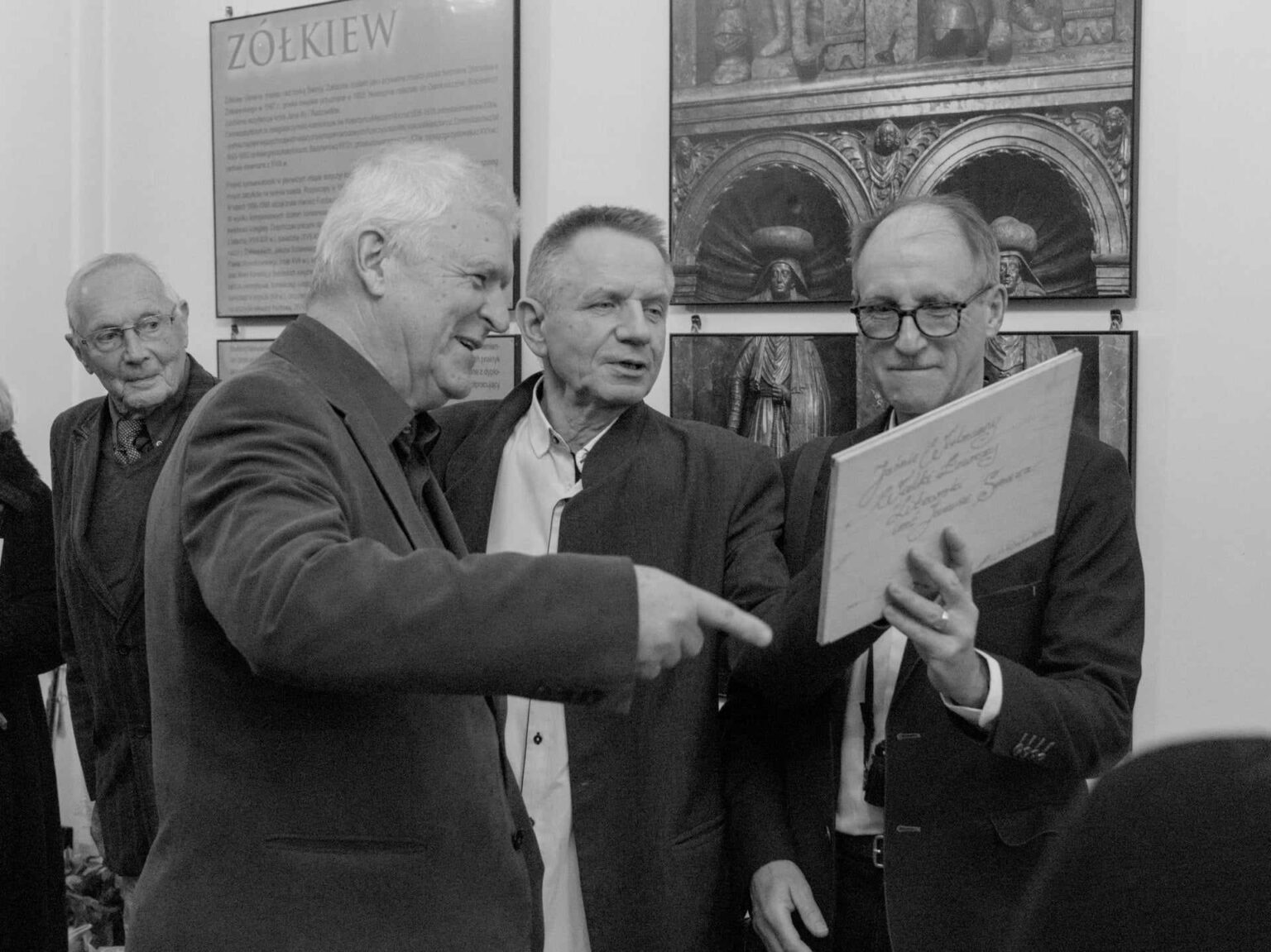 zterech starszych mężczyzn w eleganckich strojach ogląda dużą planszę z napisem. Jeden z nich uśmiecha się i wskazuje na coś na planszy. Scena ma charakter czarno-biały.