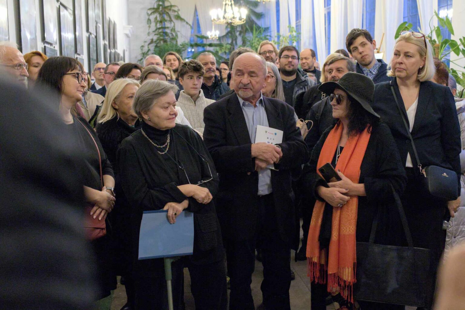 Starszy mężczyzna przemawia do tłumu ludzi w eleganckim wnętrzu. W tle widać grupę uczestników wydarzenia, dekoracyjną roślinność oraz żyrandol.