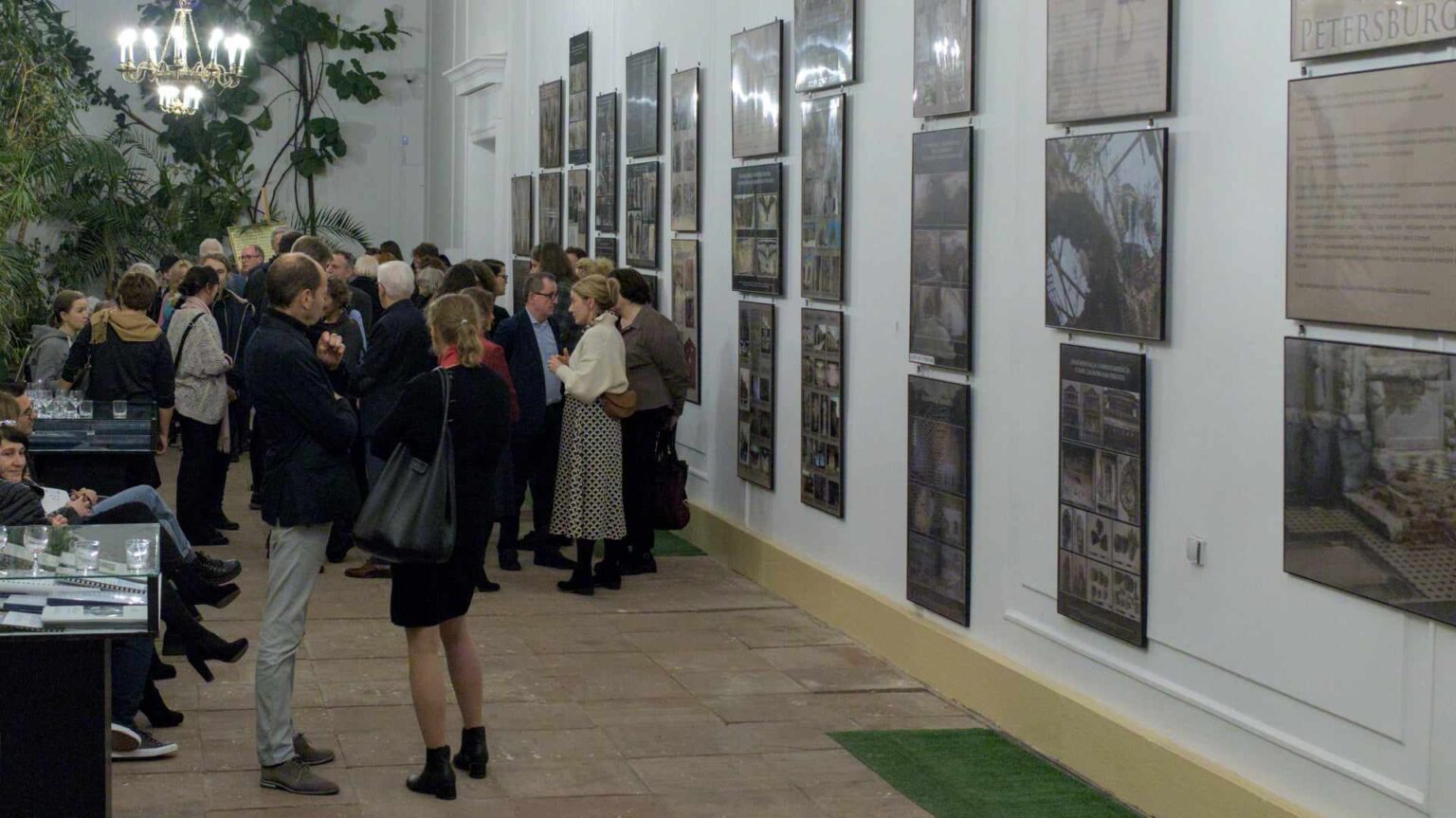 Wnętrze eleganckiej sali wystawowej z białymi ścianami, na których wiszą oprawione plansze z tekstem i zdjęciami. Grupa ludzi w różnym wieku rozmawia i ogląda wystawę. W tle widoczna jest dekoracyjna roślinność i żyrandol.