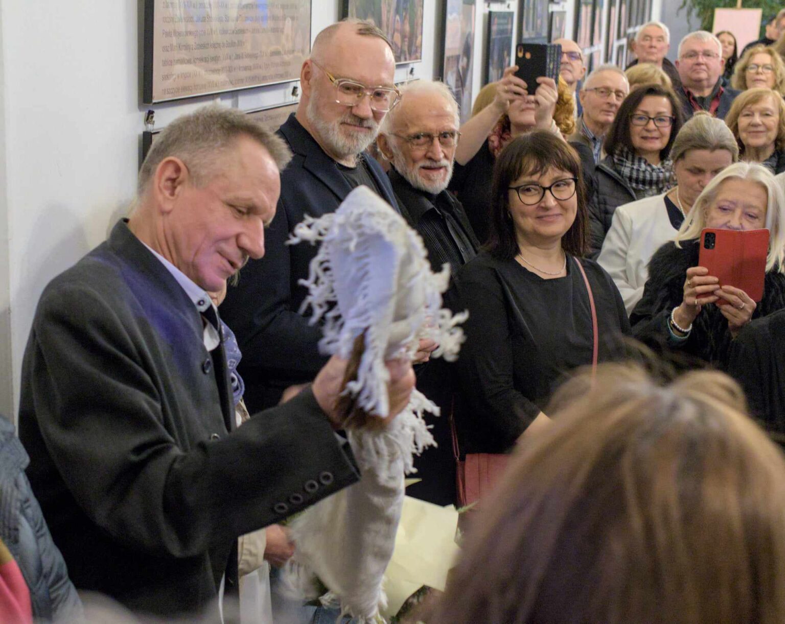 Mężczyzna w eleganckim stroju trzyma ozdobny materiał, który oglądają z uśmiechem zgromadzeni wokół niego uczestnicy wydarzenia. W tle widoczne są plansze wystawowe.