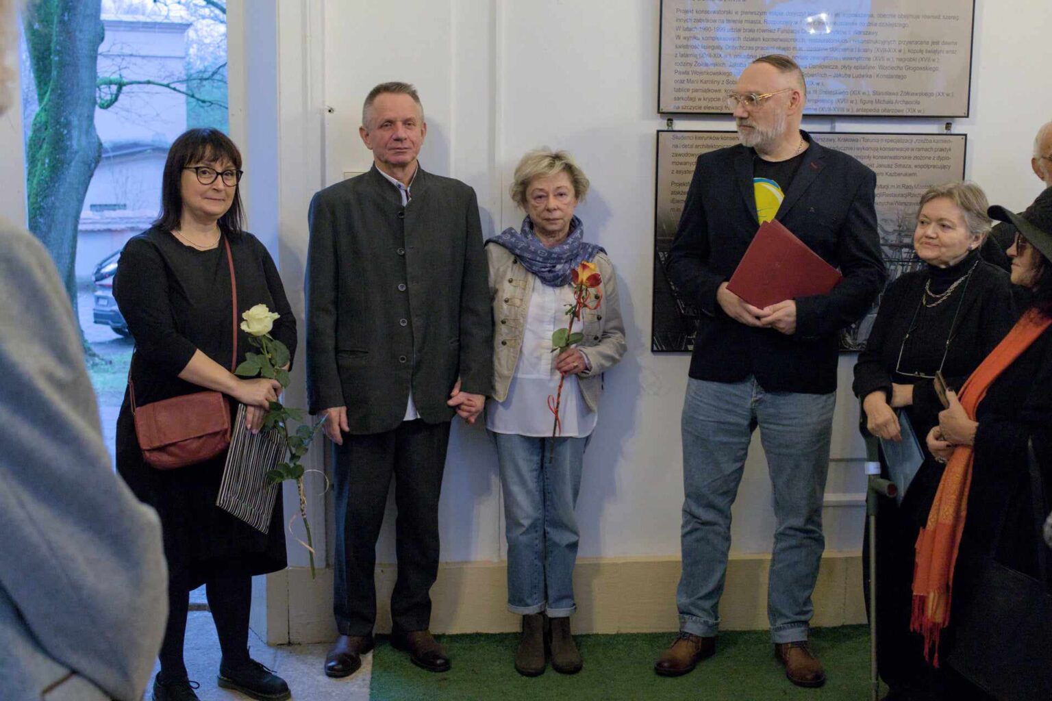 Trzy osoby stojące w grupie na tle białej ściany. Kobieta z bukietem kwiatów stoi obok mężczyzny w eleganckim stroju. Obok nich znajduje się starsza kobieta z różą. Na ścianie widoczne są plansze z tekstem.