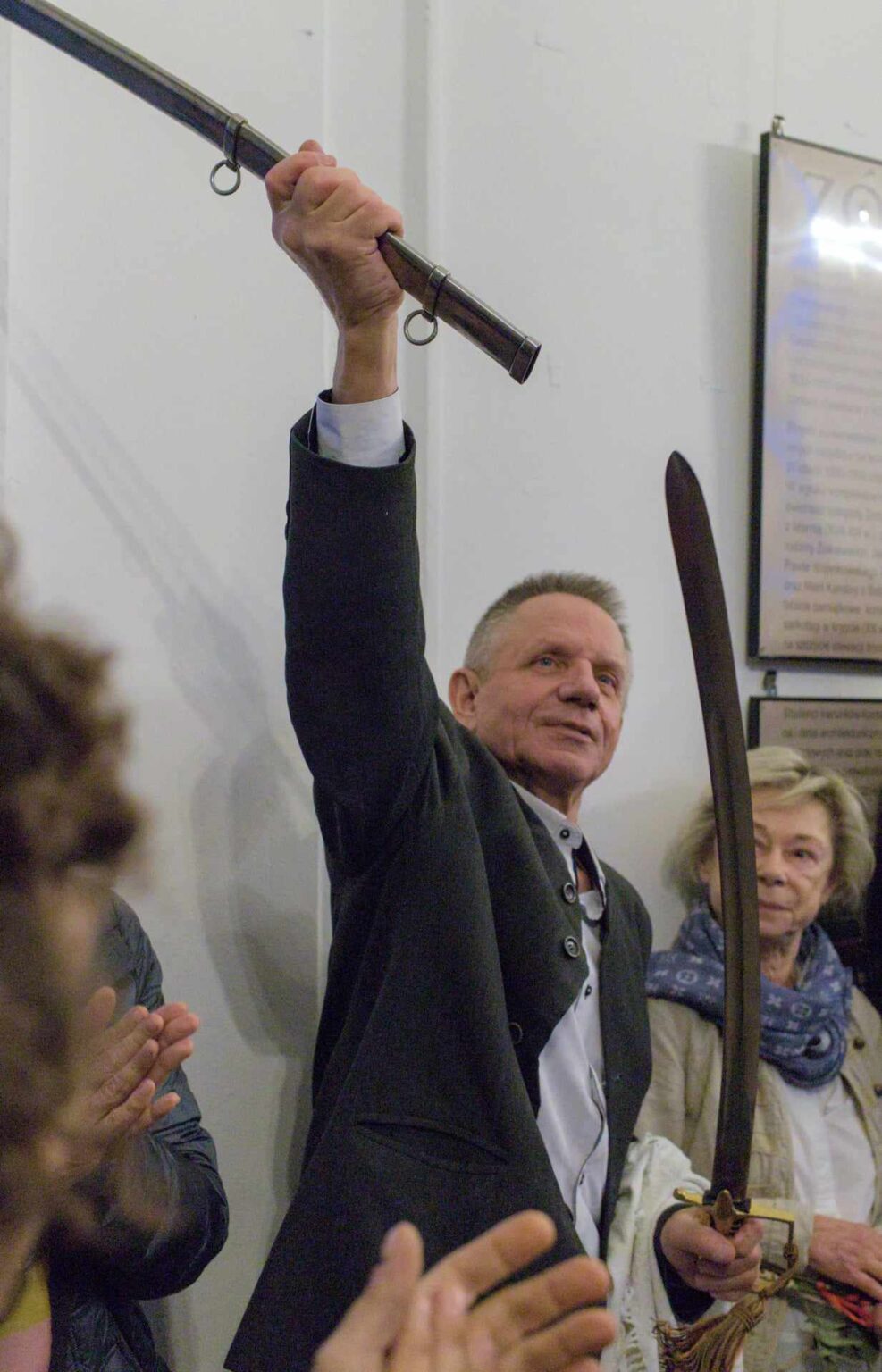 Mężczyzna z uniesioną szablą w dłoni otoczony przez grupę ludzi. Na twarzy widnieje wyraz dumy, a inni uczestnicy biją brawo. W tle widoczne są plansze wystawowe.