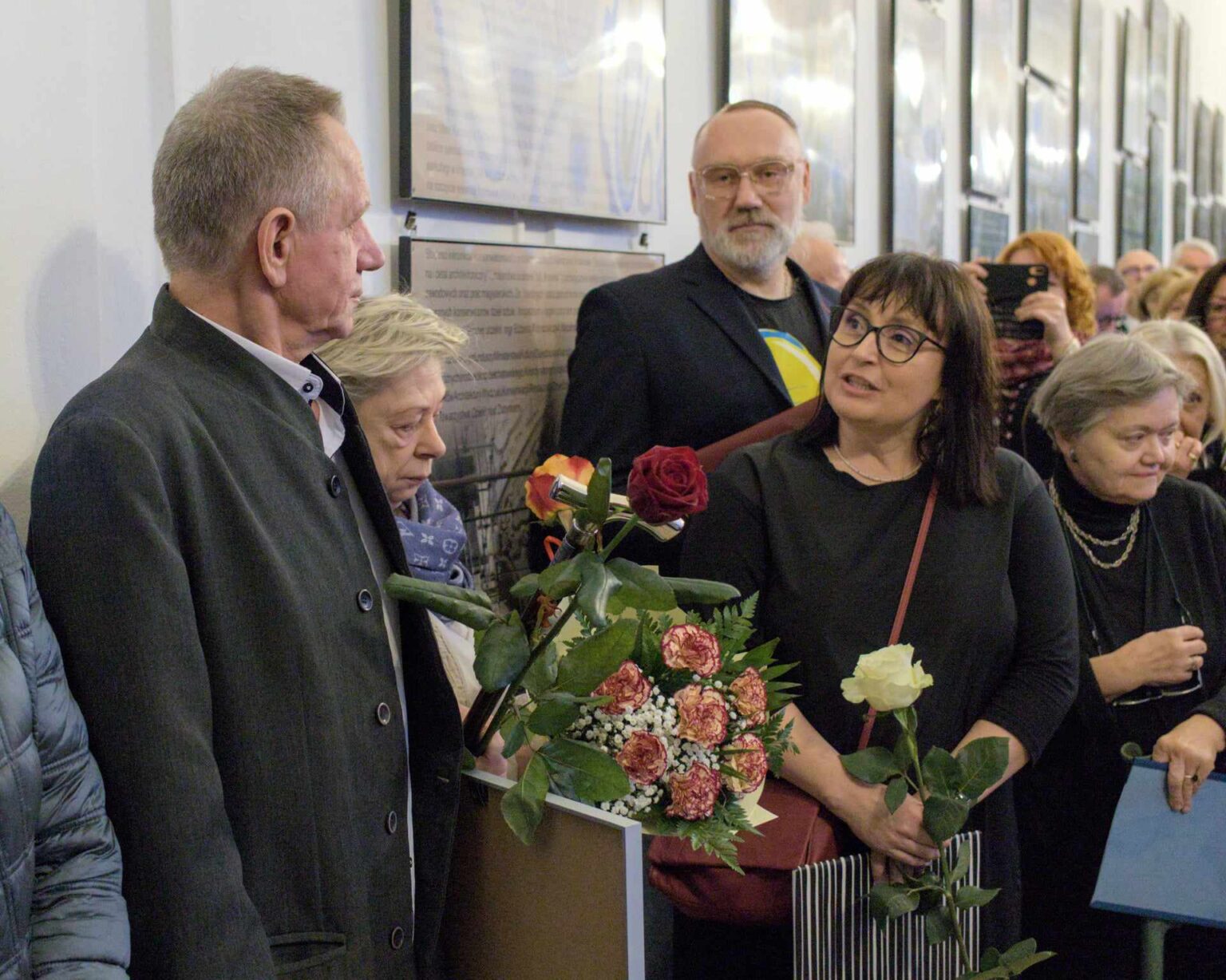 Grupa osób w różnym wieku uczestniczy w wydarzeniu kulturalnym. Na pierwszym planie kobieta z bukietem kwiatów rozmawia z innymi uczestnikami, w tle widoczne są plansze wystawowe.