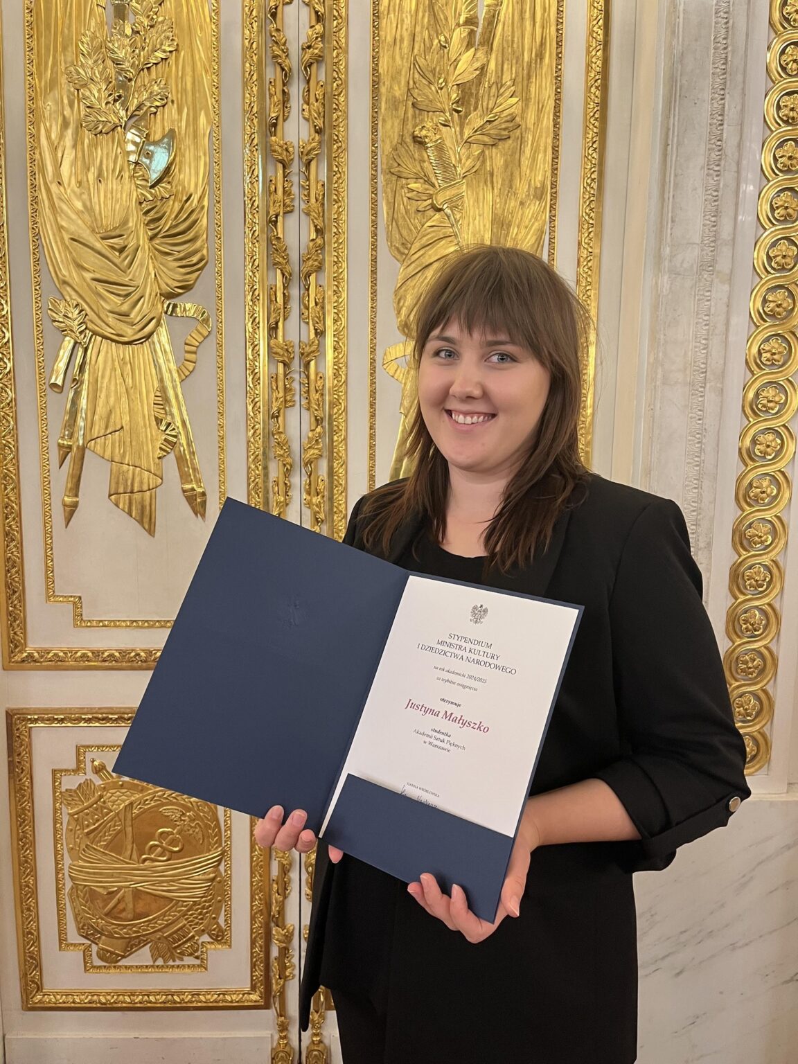 Młoda kobieta w eleganckim stroju trzyma otwarty dyplom Stypendium Ministra Kultury i Dziedzictwa Narodowego. Tło stanowi bogato zdobiona, złota ściana, co podkreśla podniosłość chwili i prestiż wyróżnienia. Kobieta uśmiecha się, prezentując dokument.