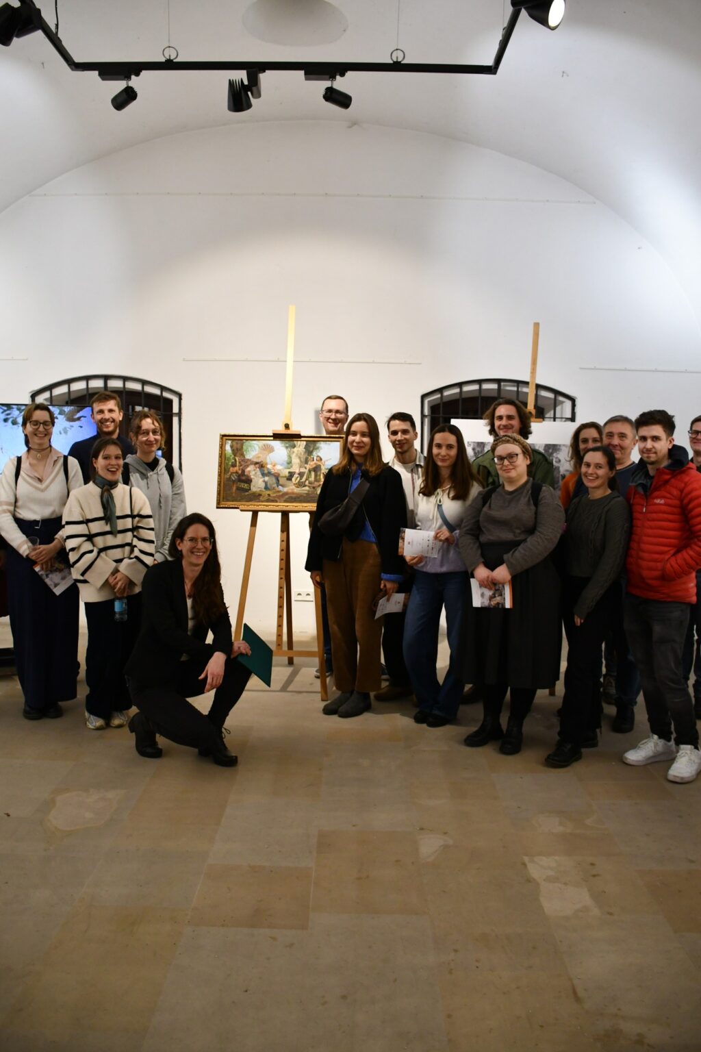 Grupa osób pozuje na tle jasnej sali wystawowej. Na środku znajduje się sztaluga z oprawionym obrazem w stylu antycznym. Wszyscy uczestnicy wyglądają na zaangażowanych i zadowolonych.