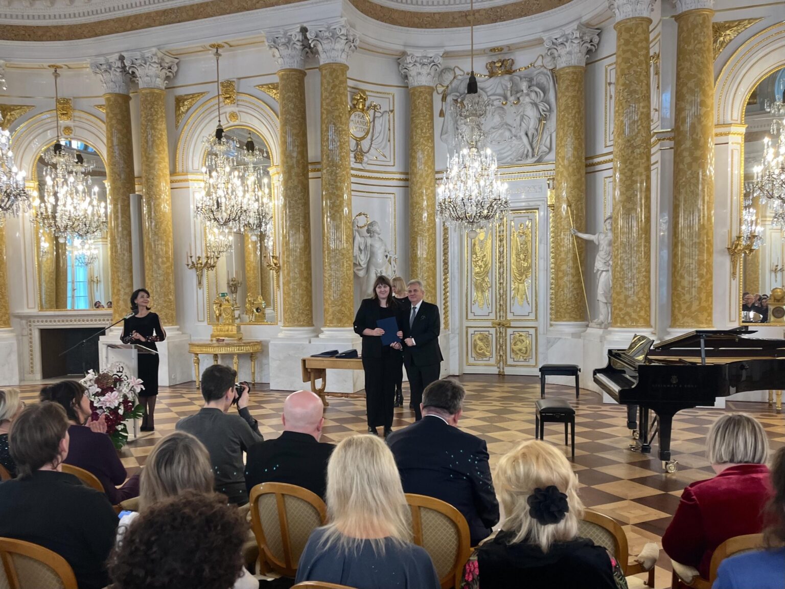 Wnętrze bogato zdobionej sali balowej z marmurowymi kolumnami i złotymi ornamentami. Na pierwszym planie widoczni są goście siedzący na krzesłach, podczas gdy na scenie w centralnej części stoi kobieta i mężczyzna. W tle błyszczące żyrandole i fortepian dodają elegancji uroczystemu charakterowi wydarzenia.