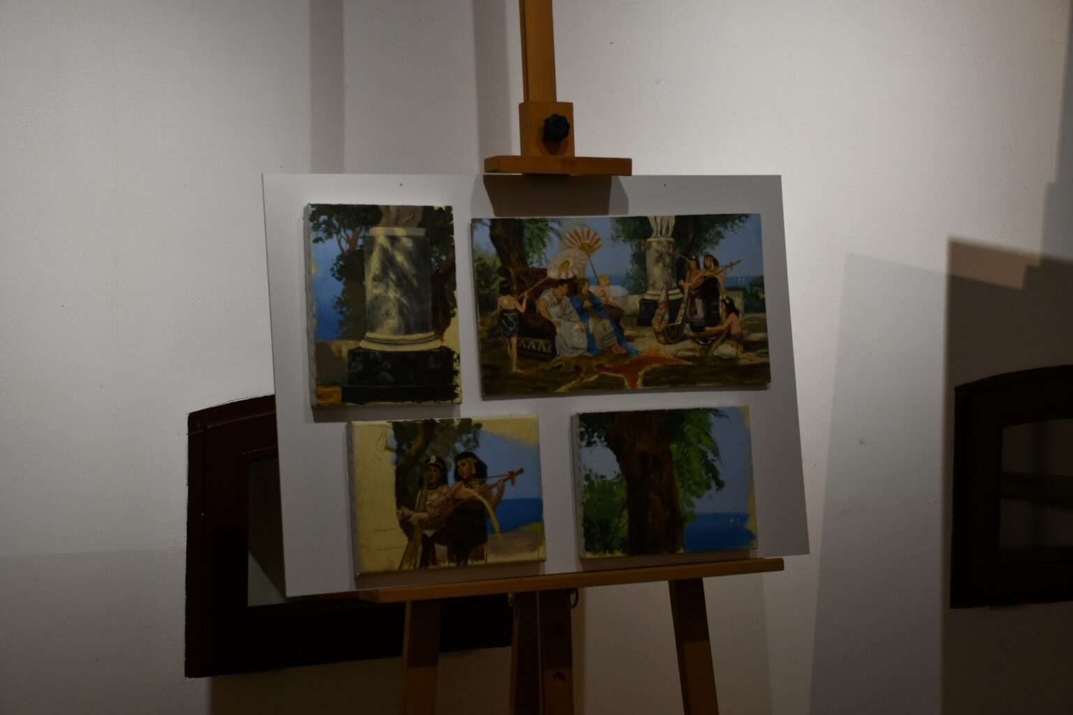 Sztaluga z białą planszą, na której znajdują się cztery szkice malarskie. Każdy z nich przedstawia różne elementy sceny plenerowej w stylu antycznym, w tym postacie i pejzaże.