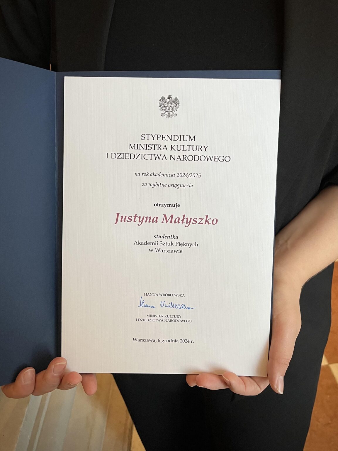 Zbliżenie na otwarty dyplom Stypendium Ministra Kultury i Dziedzictwa Narodowego. Tekst na dyplomie informuje o wyróżnieniu Justyny Małyszko, studentki Akademii Sztuk Pięknych w Warszawie, za wybitne osiągnięcia. Dokument trzymany jest w rękach osoby ubranej na czarno.