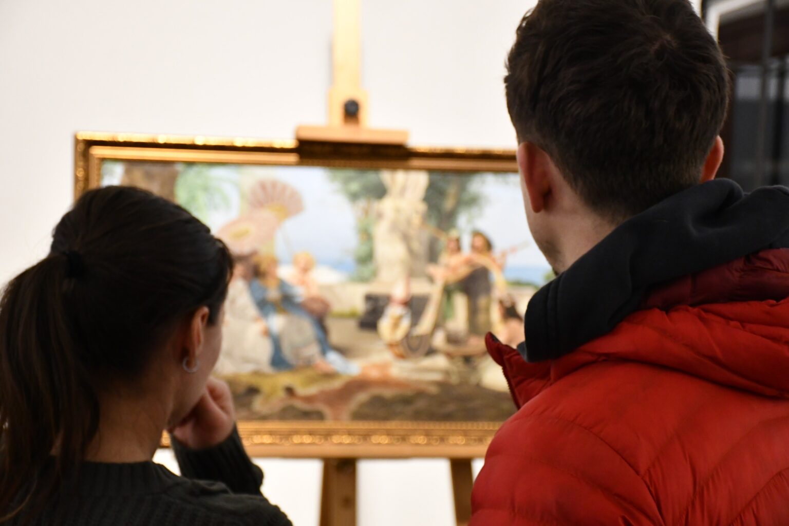 Dwoje ludzi, widocznych z tyłu, ogląda oprawiony obraz w stylu antycznym na sztaludze. Mężczyzna nosi czerwoną kurtkę, kobieta ma ciemne włosy związane w kucyk.