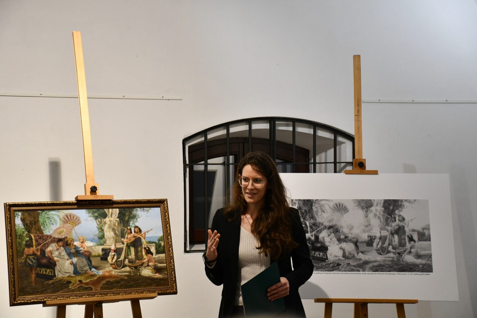 Kobieta w okularach przemawia podczas prezentacji w galerii. Na sztalugach znajdują się oprawiony obraz w kolorze oraz jego czarno-biała reprodukcja.