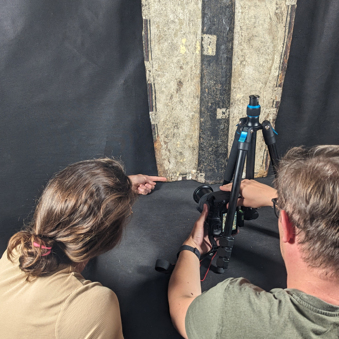 Dwie osoby dokumentujące szczegóły tarczy rycerskiej. Jedna z nich wskazuje na fragment obiektu, druga obsługuje aparat fotograficzny na statywie, przygotowując się do wykonania zdjęć.