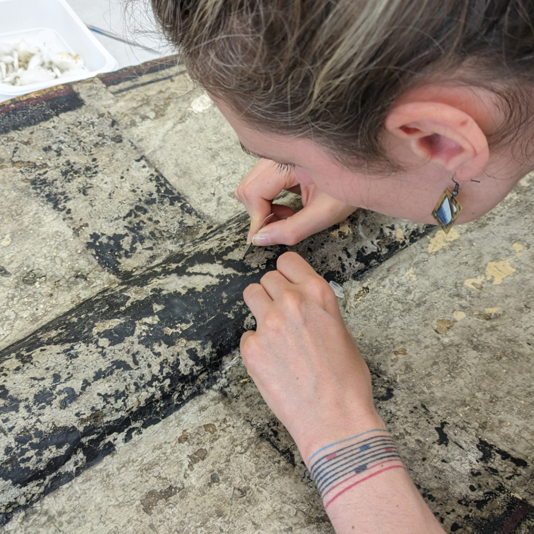 Osoba prowadząca konserwację tarczy rycerskiej. Kobieta precyzyjnie pracuje nad odnowieniem powierzchni tarczy, skupiając się na detalu malarskim.