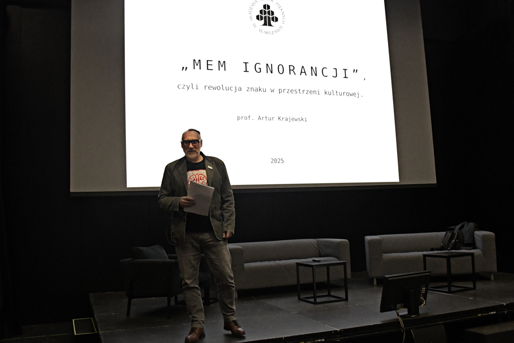 Mężczyzna w okularach stoi na scenie przed ekranem projekcyjnym. Na ekranie widnieje napis: „MEM IGNORANCJI, czyli rewolucja znaku w przestrzeni kulturowej”, podpisany „prof. Artur Krajewski, 2025”. Scena jest wyposażona w minimalistyczne meble: dwie kanapy i małe stoliki.