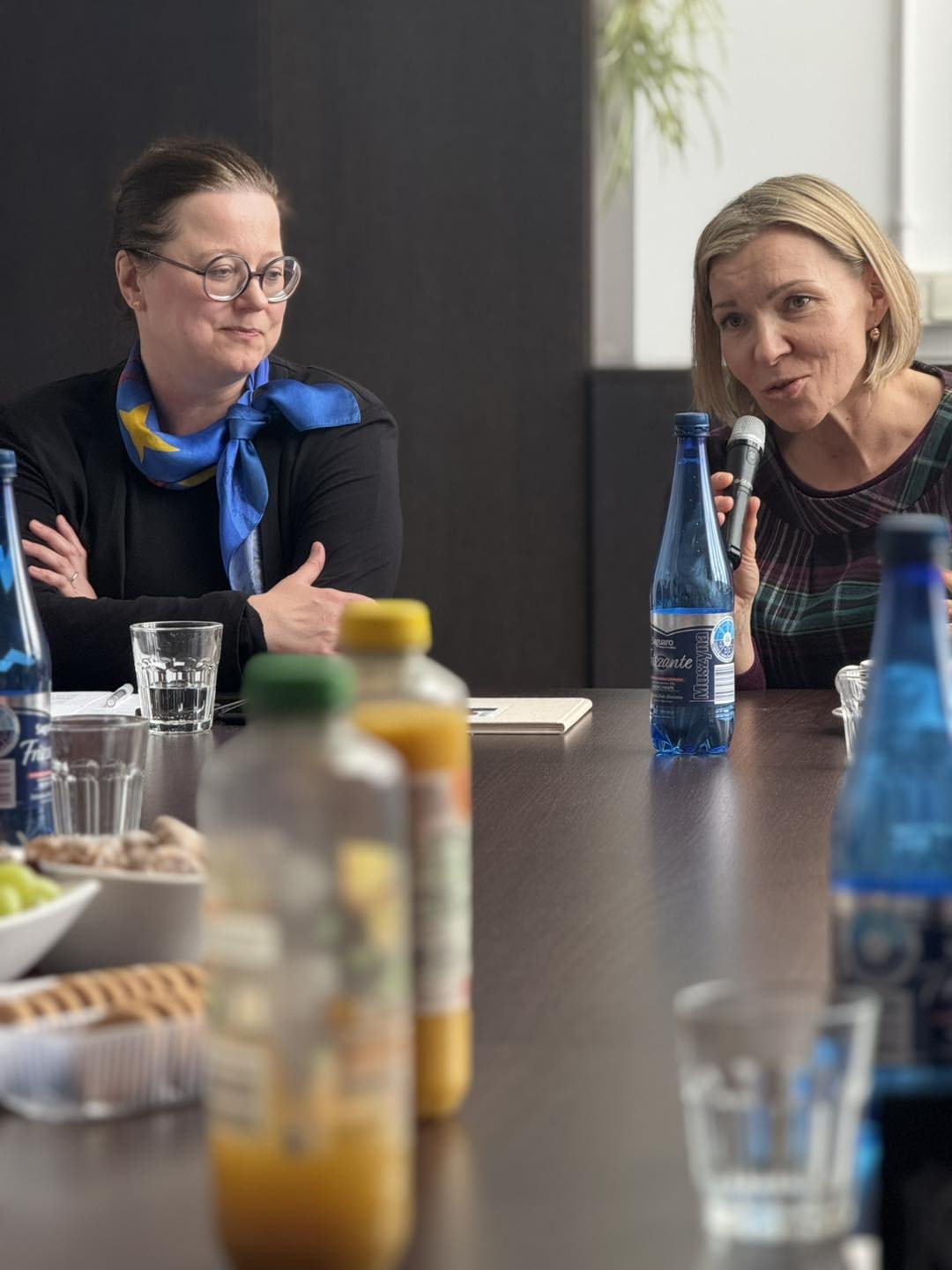 Dwie kobiety przy stole, jedna mówi do mikrofonu.