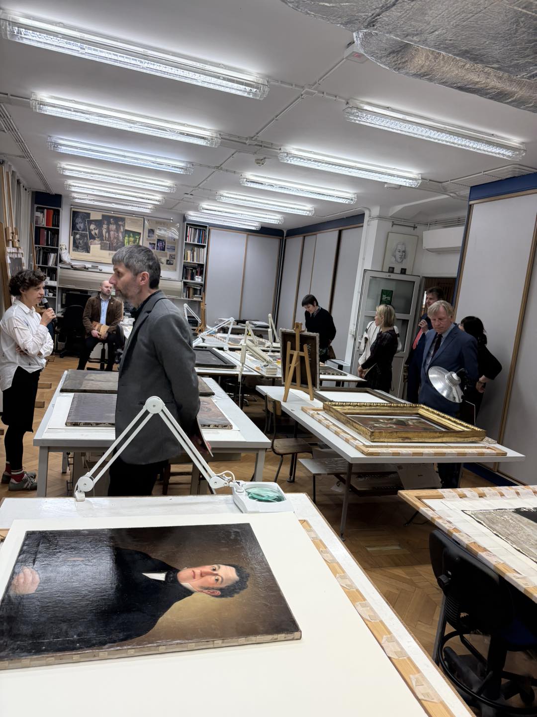 Grupa ludzi stoi pomiędzy stołami na których leżą zabytkowe obrazy