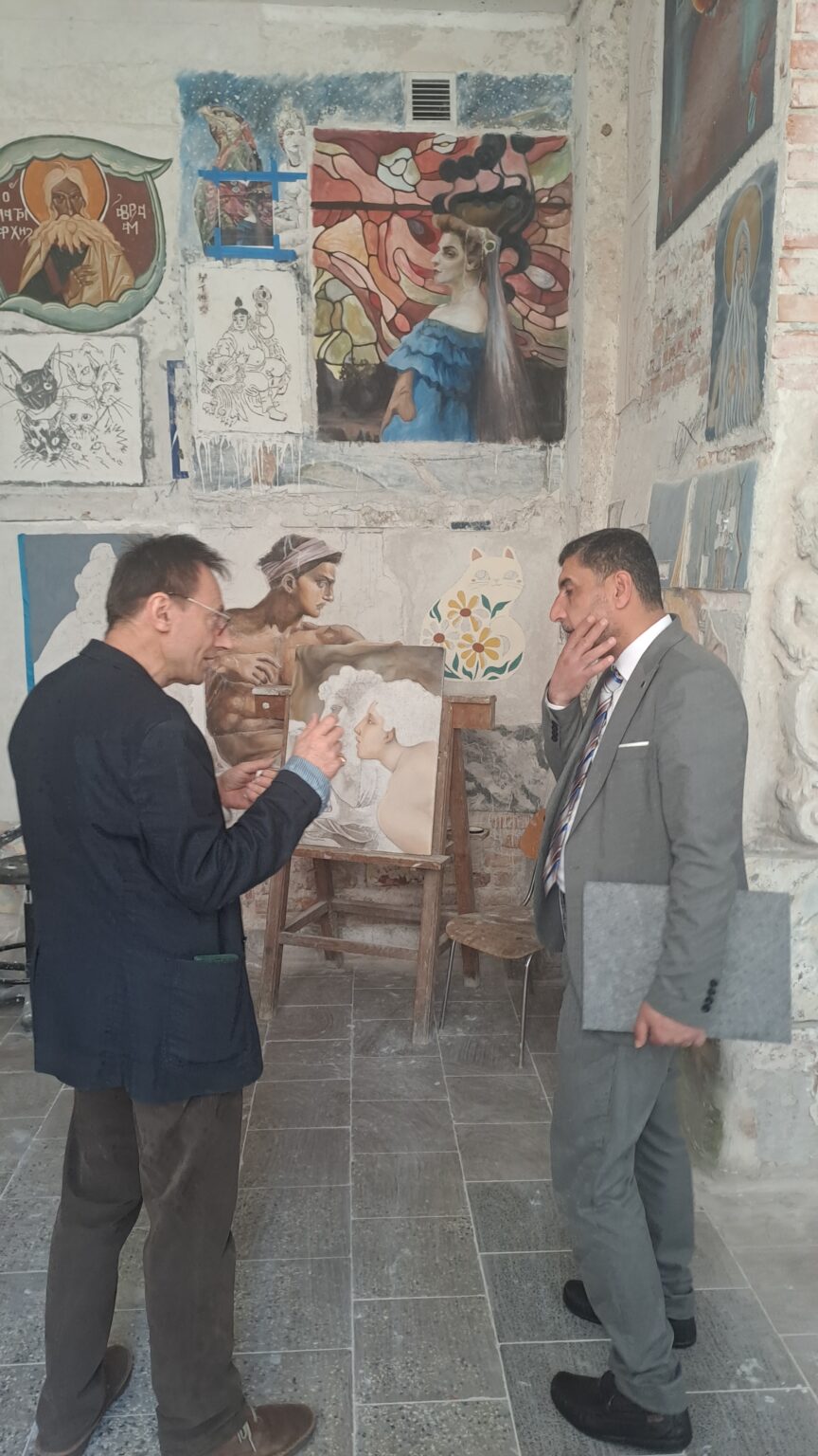 Dwóch mężczyzn rozmawia w pracowni artystycznej, otoczeni szkicami i obrazami na ścianie. Jeden z nich, w granatowej marynarce, gestykuluje ręką, jakby tłumaczył coś na temat prezentowanej pracy malarskiej – studium dwóch kobiecych profili. Drugi mężczyzna, w szarym garniturze, słucha z zainteresowaniem, trzymając w ręku teczkę i dotykając brody w zamyśleniu.