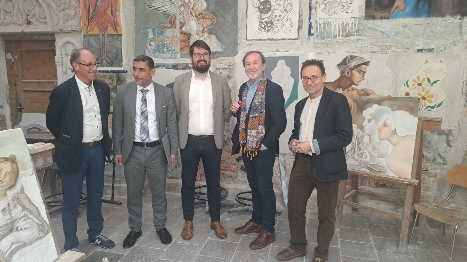 Pięciu mężczyzn stoi w rzędzie na tle artystycznie zaaranżowanej ściany z malowidłami i szkicami. Po lewej stronie widoczna jest sztaluga z obrazem kobiecego aktu, a w centralnej części ściany – realistycznie namalowana postać młodego mężczyzny oraz rysunkowy kot z kwiatowym wzorem. Uczestnicy wyglądają na zadowolonych, niektórzy uśmiechają się lub spoglądają w bok.