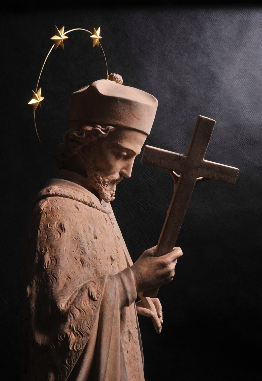 Rzeźba świętego (najpewniej św. Jana Nepomucena) trzymającego krzyż. Postać z aureolą utworzoną z metalowego pierścienia z pięcioma złotymi gwiazdami, ubrana w szatę z wyraźną fakturą i brodą.