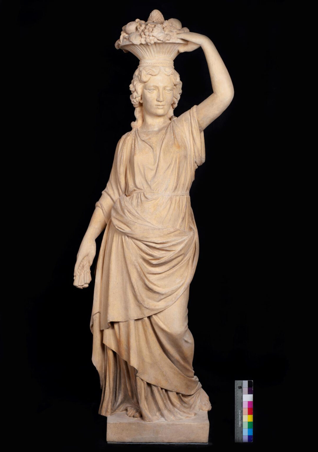 Klasycystyczna rzeźba kobiety z koszem owoców na głowie. Postać ubrana w długą, fałdzistą szatę, stoi w kontrapoście na prostokątnym postumencie, z jedną ręką opuszczoną, drugą podtrzymującą kosz.
